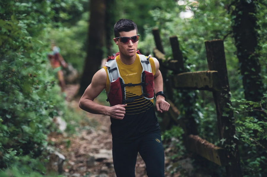 Man trail running in sports sunglasses, yellow top, and hydration vest on forest path